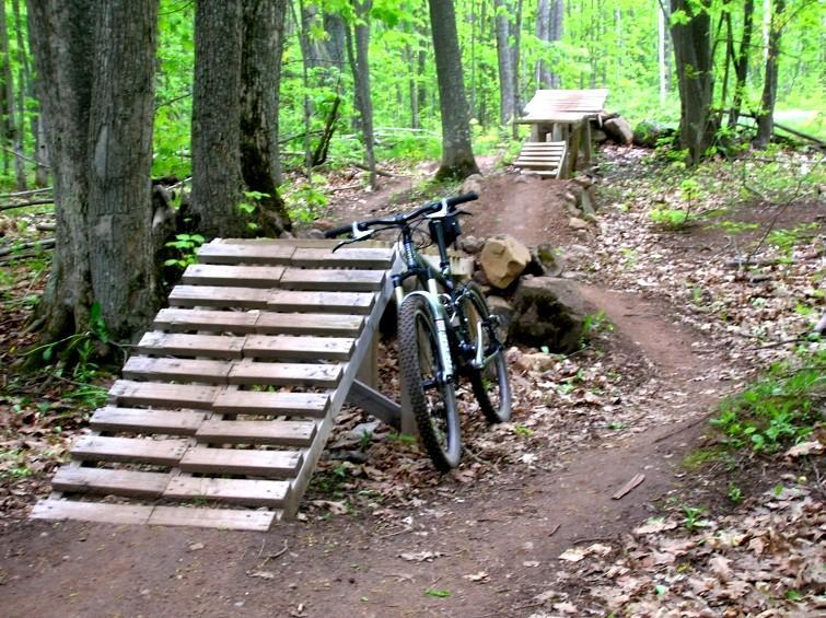 A mountain bike is parked next to wooden ramps on a dirt trail surrounded by trees and greenery. The scene captures a secluded forest area designed for biking, featuring two ramps positioned at different heights. The ground is covered with leaves, indicating it is a natural outdoor setting. Michigan Tech Trails mountain bike trail.