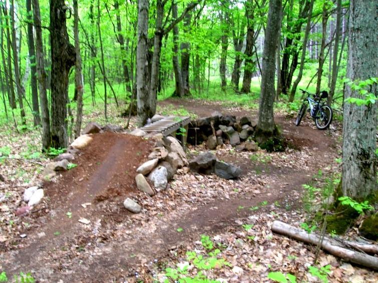 A forested area featuring a dirt bike trail with a wooden ramp and rock edges, surrounded by green trees and scattered leaves. A mountain bike is parked nearby on the trail. Michigan Tech Trails mountain bike trail.
