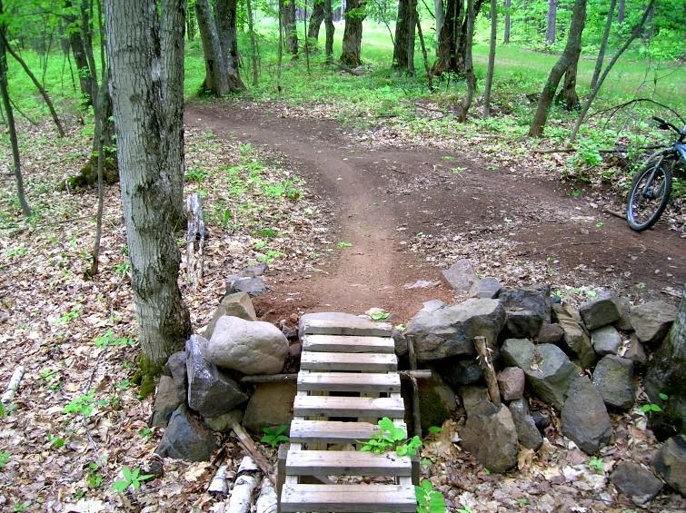 A narrow wooden bridge made of slats crosses over a small gap, surrounded by rocks, in a lush green forest. A winding dirt path leads away from the bridge, and a mountain bike is parked nearby. The scene is filled with trees and foliage, creating a tranquil outdoor setting. Michigan Tech Trails mountain bike trail.