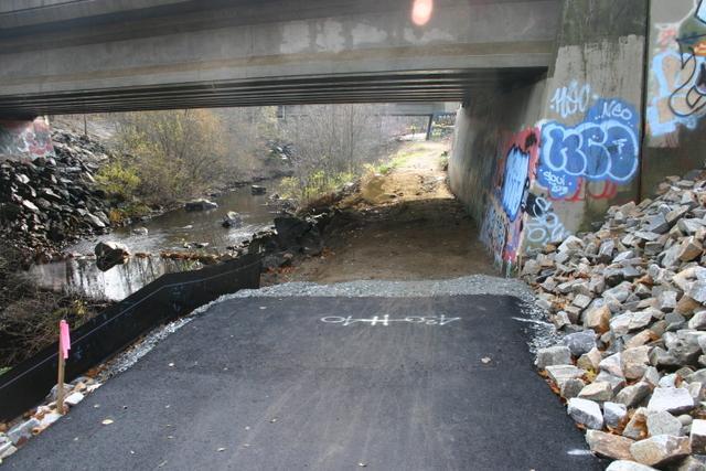 A view of a small trail that leads under a concrete overpass, with a creek visible on the left side. The ground is newly paved with a dark surface, and there are large rocks piled on the right. The concrete walls of the overpass show various graffiti marks, and the surrounding area features sparse trees and vegetation. Trumbull Rail Trail mountain bike trail.