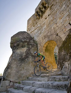 A mountain biker riding down stone steps near a historic archway, surrounded by large rocks and ancient stone walls. The scene is set against a clear sky, highlighting the cyclist's action.
