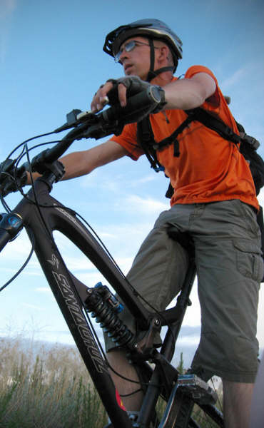 A person wearing an orange shirt and helmet is riding a mountain bike, positioned in a natural outdoor setting with a clear sky in the background. The rider appears focused and poised, with a backpack on, showcasing a dynamic cycling posture.