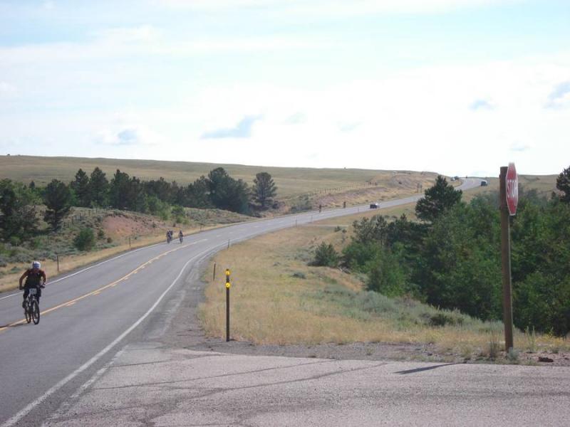 A two-lane road curves through a rural landscape, with cyclists riding along the left side. In the foreground, a stop sign stands next to the road, while green trees and grass line the sides. The road continues into the distance, flanked by open fields and a clear blue sky. Laramie Enduro mountain bike trail.