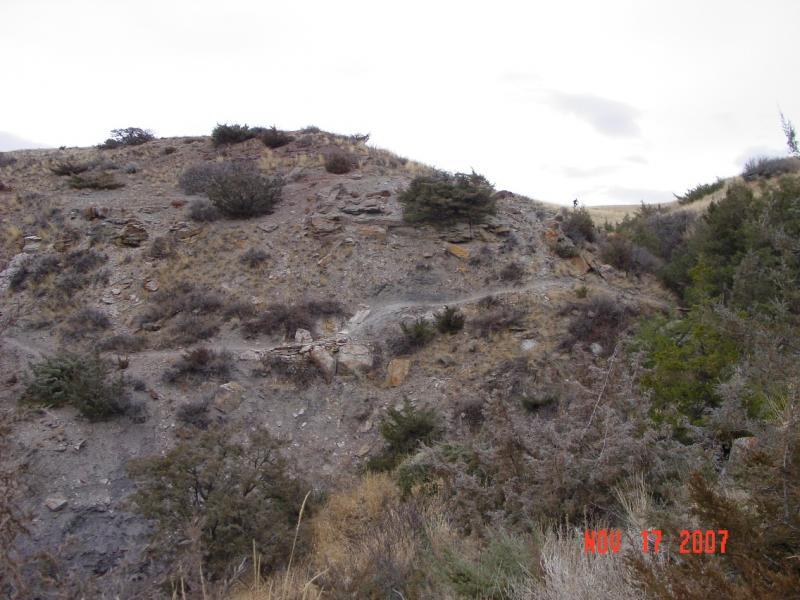 A rugged hillside covered with sparse vegetation and rocky terrain, set against a cloudy sky. A winding path can be seen leading up the slope, with shrubs and scattered trees in the foreground. The image is dated November 17, 2007. River's Edge mountain bike trail.