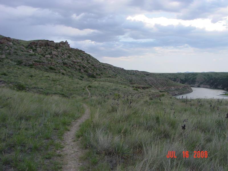 A scenic landscape featuring a winding dirt path through grassy terrain, leading towards a river bordered by rocky hills under a cloudy sky. The image hints at a tranquil, natural setting with lush vegetation and distant hills. North Shore mountain bike trail.