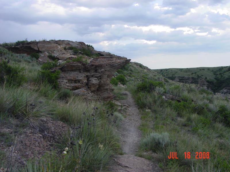 A scenic view of a hiking trail winding through a rocky landscape, featuring a distinct rock formation on the left surrounded by green vegetation and hilly terrain under a cloudy sky. North Shore mountain bike trail.
