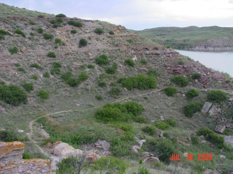 A scenic view of a grassy hillside with rocky outcrops and sparse vegetation leading down to a body of water. The landscape is dotted with shrubs and small trees, with gentle slopes and a cloudy sky in the background. The date in the lower right corner indicates July 16, 2008. North Shore mountain bike trail.