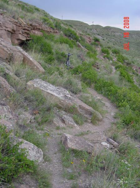A narrow, winding dirt path leads through lush green grass and rocky terrain on a hillside, with a bicycle leaning against a rock in the background. The scene is captured under a cloudy sky, suggesting an outdoor hiking or biking trail. North Shore mountain bike trail.