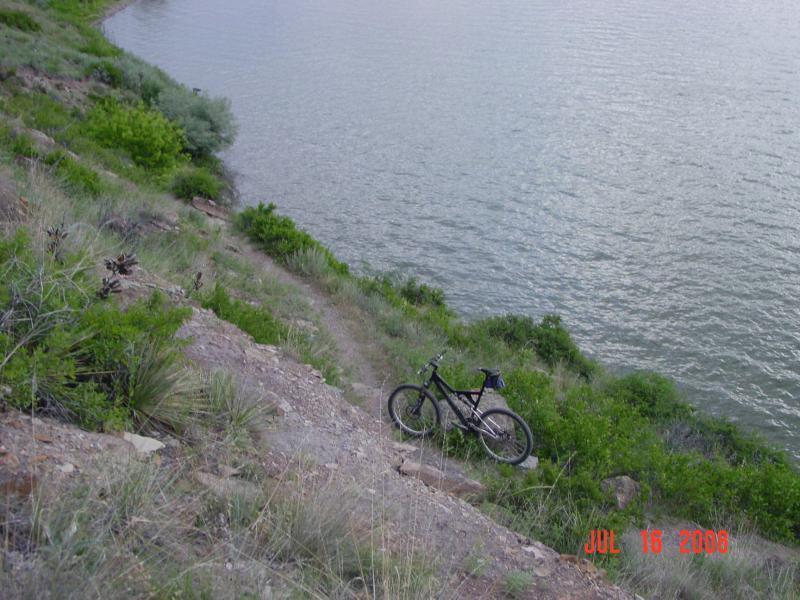 A black mountain bike is resting on a gravelly slope near a calm body of water, surrounded by lush green vegetation. The image captures the landscape of the area, showcasing the incline leading down to the water's edge. North Shore mountain bike trail.