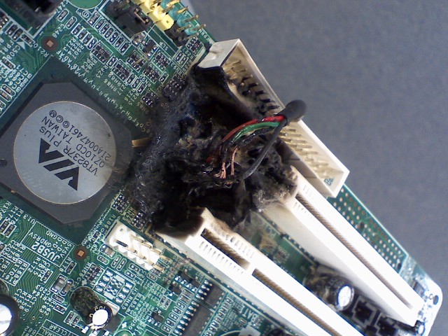 Close-up view of a damaged computer motherboard showing burn marks and melted components. Wires appear singed, and blackened residue is present around the area of damage.