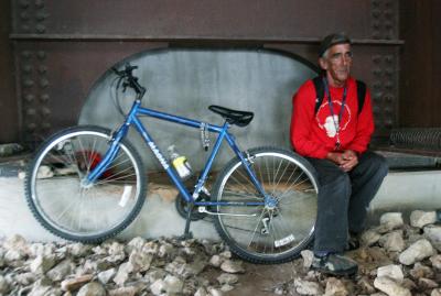 A man sitting on a rocky surface under a bridge, wearing a red sweatshirt and gray pants, with a blue bicycle leaning against a wall beside him.