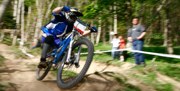 A mountain biker in a blue outfit leans into a turn on a dirt trail, showcasing dynamic movement on a rocky path. In the background, spectators watch from the sidelines amidst a forested setting.