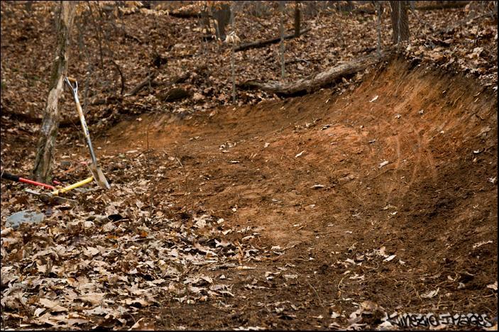 A dirt biking trail in a wooded area, featuring a smooth, dirt surface with scattered fallen leaves. A shovel and a rake are leaned against a tree in the foreground, indicating recent maintenance or trail work. Wild Turkey mountain bike trail.