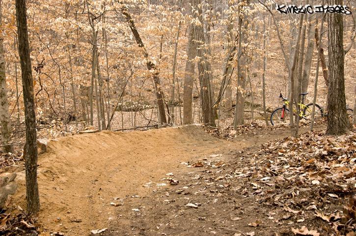 A dirt bike ramp surrounded by trees in an autumn landscape, with fallen leaves covering the ground. A yellow mountain bike is positioned nearby, suggesting an outdoor recreational area for biking and trail riding. Wild Turkey mountain bike trail.