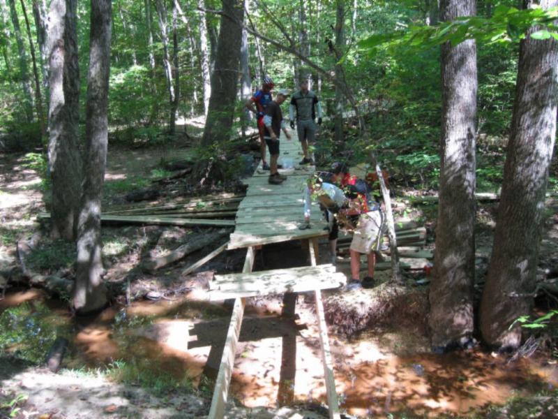 A group of people working on a wooden bridge in a forested area, surrounded by trees and a small creek. Some individuals are standing on the bridge while others are gathered nearby, with tools and materials visible in the scene. The area appears to be a nature trail or park setting. Wild Turkey mountain bike trail.