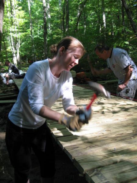 A woman in a white long-sleeve shirt is using a hammer on a wooden board as she works on a construction project in a wooded area, with another person visible in the background. Sunlight filters through the trees, highlighting the greenery. Wild Turkey mountain bike trail.