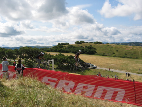 Two mountain bikers are seen in mid-air, jumping over a barrier with a logo in the foreground. A grassy hillside and spectators in the background suggest an outdoor biking event, with rolling hills and a cloudy sky visible.