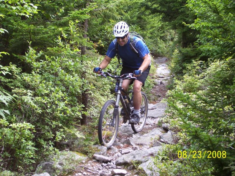 A man riding a mountain bike on a rocky trail surrounded by lush green trees. He is wearing a helmet, blue shirt, and shorts, and appears to be navigating the uneven terrain. Spruce Knob Loop mountain bike trail.