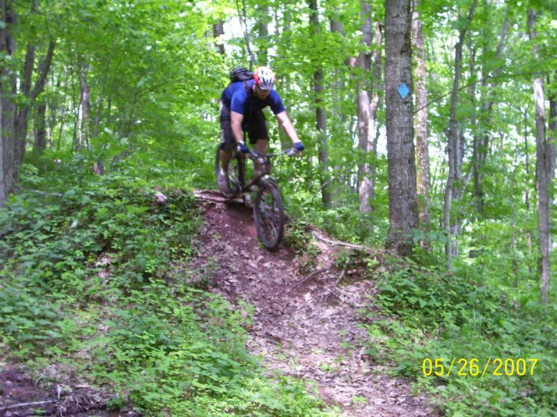 A mountain biker navigates a dirt trail in a lush green forest, riding down a slight incline with concentration. The cyclist wears a helmet and sports gear, and the surrounding trees and foliage create a vibrant, natural backdrop. The scene captures the thrill of off-road biking in a scenic outdoor environment. Props Run Trail mountain bike trail.