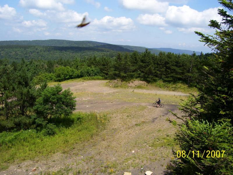 A scenic view of a forested landscape with a clear blue sky and fluffy white clouds. In the foreground, a dirt path leads through a clearing, where a person is riding a bicycle. Lush green trees line the edges of the clearing, with rolling hills visible in the background. The date in the bottom right corner indicates the image was taken on August 11, 2007. Props Run Trail mountain bike trail.