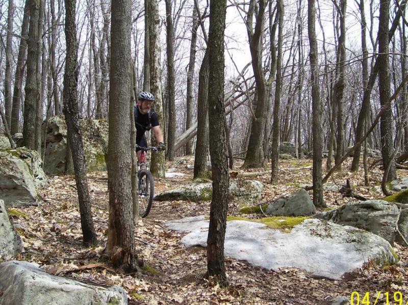 A mountain biker navigating a rocky trail through a forest with bare trees and scattered leaves, surrounded by large boulders and a natural landscape. The scene captures the outdoorsy spirit of mountain biking in a rugged environment. Big Bear Lake Trail Center mountain bike trail.