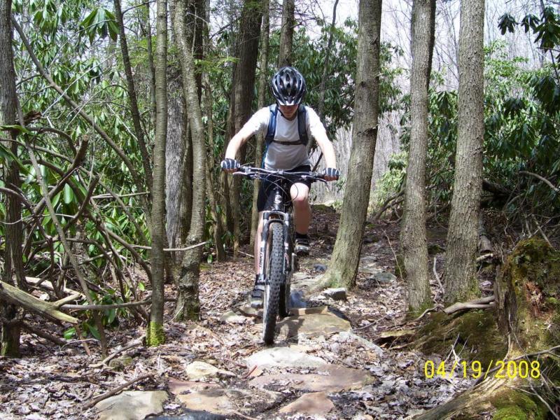 A person riding a mountain bike on a rocky trail through a wooded area, surrounded by trees and greenery. The rider is wearing a helmet and a backpack, focused on navigating the terrain. Big Bear Lake Trail Center mountain bike trail.