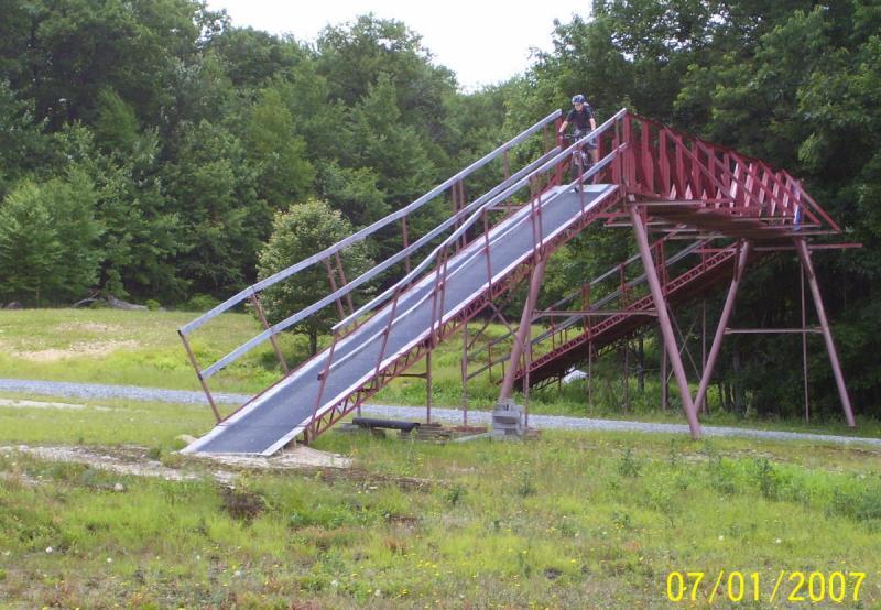 A large red metal ramp structure in a grassy area, with a person on a bicycle riding down the sloped surface. Trees are visible in the background, and the ground is a mix of grass and gravel. The image appears to be taken in a recreational or outdoor setting. Big Bear Lake Trail Center mountain bike trail.