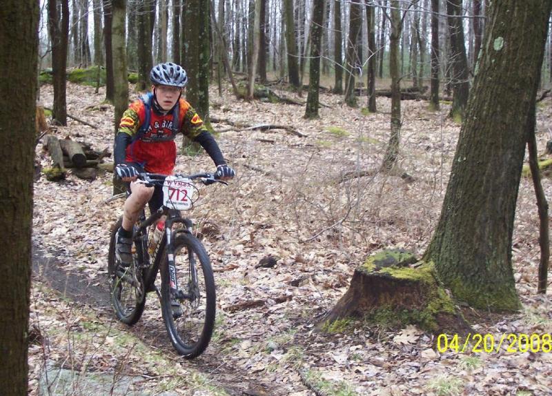 A young cyclist in a colorful jersey and helmet rides a mountain bike along a dirt path in a wooded area. The ground is covered with fallen leaves, and the trees are bare, indicating early spring. A large tree stump is visible on the right side of the image. The biker is focused and appears to be navigating the trail. Big Bear Lake Trail Center mountain bike trail.