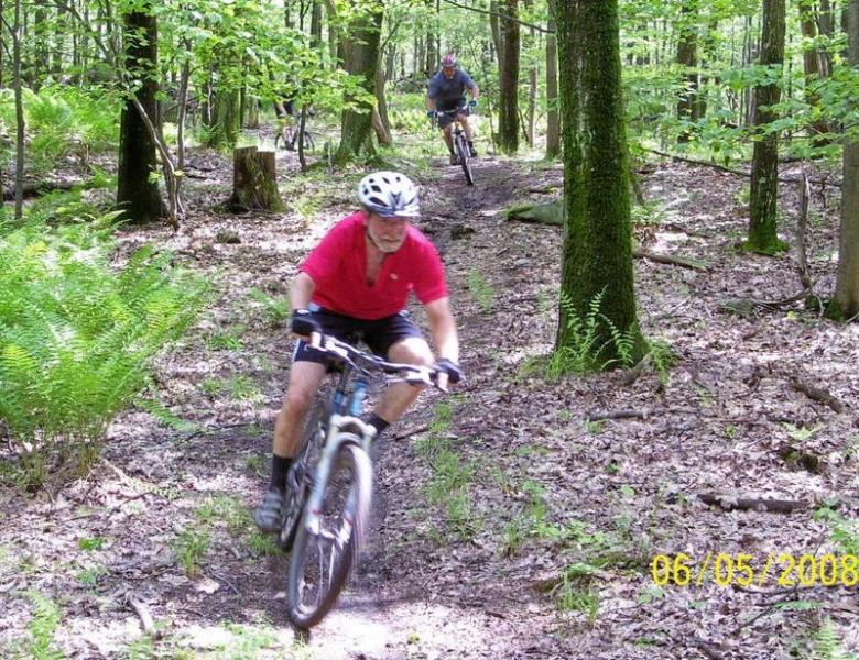 Two mountain bikers navigating a wooded trail. The rider in the foreground, wearing a red shirt and helmet, is leaning forward as they ride downhill. The second rider is visible in the background, following on a winding path through the trees. The landscape includes lush greenery and ferns, with sunlight filtering through the leaves. Big Bear Lake Trail Center mountain bike trail.