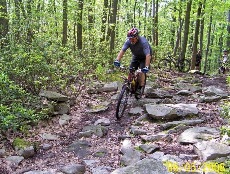 A mountain biker navigates a rocky trail in a lush green forest, with trees surrounding the path. The rider is wearing a helmet and riding gear, focused on maintaining balance over the uneven terrain. In the background, two other cyclists can be seen resting on their bikes. Big Bear Lake Trail Center mountain bike trail.