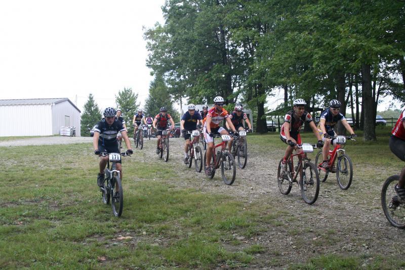 A group of mountain bikers riding on a dirt path through a grassy area, surrounded by trees. The cyclists, wearing helmets and racing jerseys, are in a line, with some displaying their race numbers. A building can be seen in the background. Big Bear Lake Trail Center mountain bike trail.