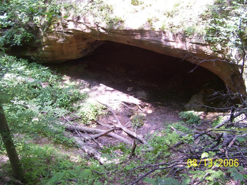 A natural rock overhang surrounded by dense green foliage, with scattered branches and underbrush on the forest floor. The opening reveals a shadowed interior, suggesting a cave or shelter. Sunlight filters through the trees, illuminating parts of the landscape. Archers Fork Loop Trail mountain bike trail.