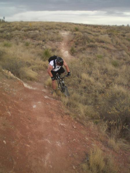 A mountain biker navigating a dirt trail in a grassy, open landscape. The biker is wearing a helmet and a black and white jersey, leaning forward as they maneuver around a bend in the path. The terrain is rugged, with sparse vegetation and a cloudy sky above. Skidmarks mountain bike trail.