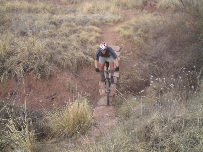 A mountain biker riding over a rocky path in a grassy terrain, surrounded by tall grass and earthy tones, with a slight incline ahead. The cyclist is wearing a helmet and riding posture suggests active movement along the trail. Skidmarks mountain bike trail.