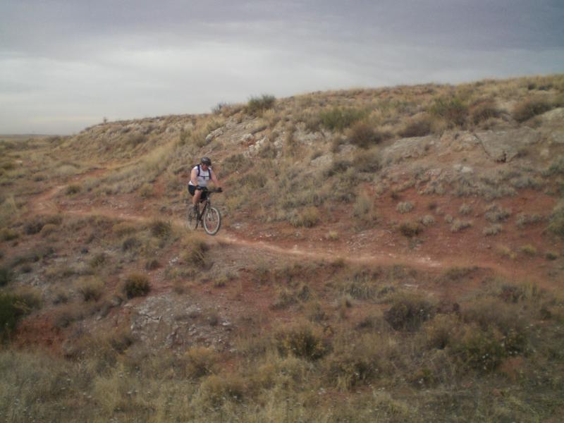A person riding a mountain bike on a dirt trail through a grassy, hilly landscape under an overcast sky. Skidmarks mountain bike trail.