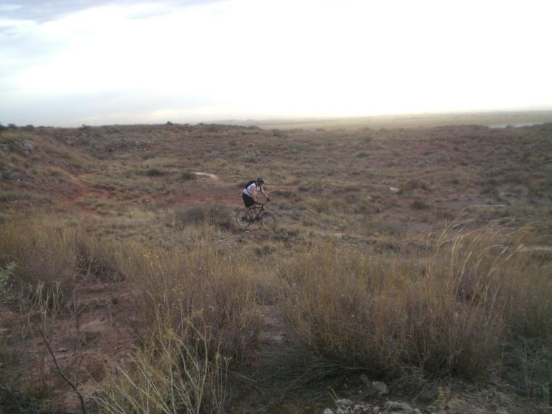 A mountain biker rides through a rugged, grassy landscape under a cloudy sky, with distant hills visible in the background. Skidmarks mountain bike trail.