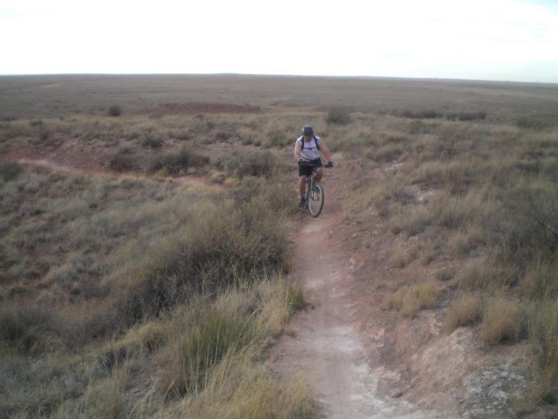A cyclist riding a mountain bike on a narrow dirt trail surrounded by grassy terrain and open landscapes under a cloudy sky. Skidmarks mountain bike trail.