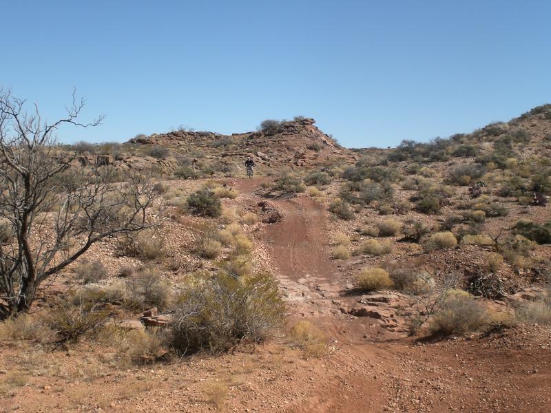 A dusty trail winds through a rugged, arid landscape with scattered shrubs and sparse vegetation. In the background, a rocky hillside rises under a clear blue sky. A lone figure is seen walking along the trail, emphasizing the remote and natural setting. Haystack Mountain mountain bike trail.