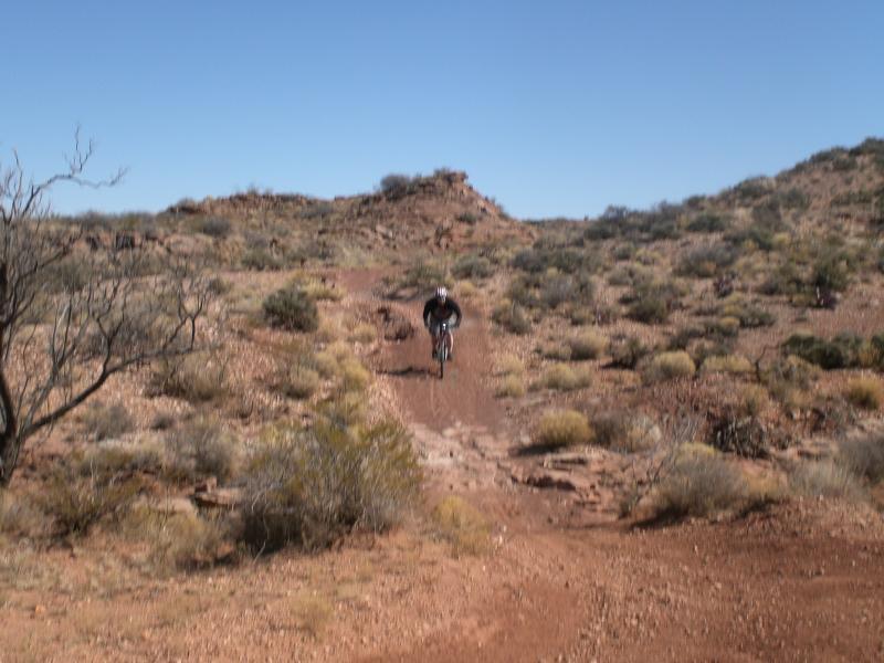 A mountain biker rides along a dirt trail in a rugged, arid landscape with sparse vegetation and rocky hills under a clear blue sky. Haystack Mountain mountain bike trail.