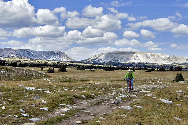 A mountain biker rides along a rocky path in a wide-open grassy landscape, with large mountains in the background and a partly cloudy sky overhead. Libby Flats mountain bike trail.