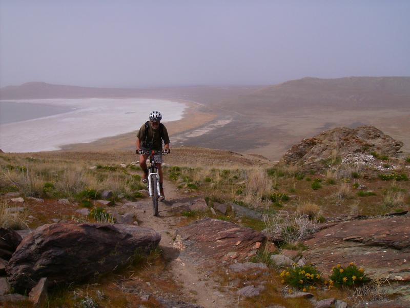 A mountain biker navigating a rocky trail on a hillside, overlooking a vast, open landscape with a beach and water in the background. The scene is slightly hazy, suggesting a misty atmosphere, with patches of green grass and scattered rocks in the foreground. Antelope Island mountain bike trail.