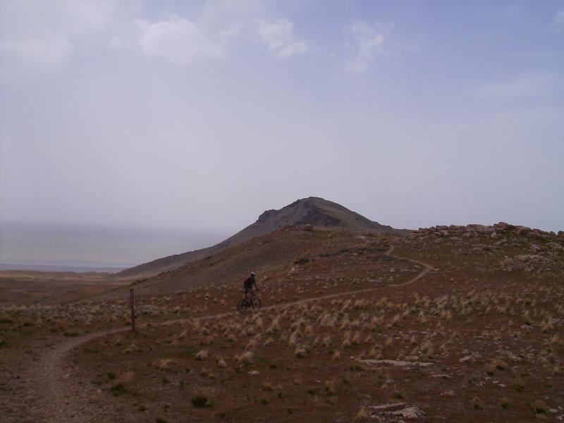 A cyclist riding along a dirt path in a remote, hilly landscape under a cloudy sky. A mountain rises in the background, surrounded by sparse vegetation. Antelope Island mountain bike trail.