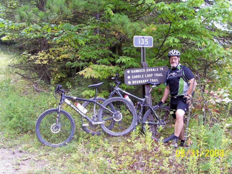 A man in cycling attire stands next to two mountain bikes at a trail sign indicating directions for Bannock Shoals Trail, Saddle Loop Trail, and Boundary Trail. The background features green foliage, suggesting a forested area. The image is dated August 31, 2008. Bear Pen (modified) Loop mountain bike trail.