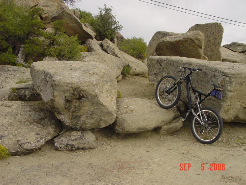 A mountain bike leaning against large boulders on a dirt trail, surrounded by greenery. The image features rocky terrain and a cloudy sky in the background. Zimmerman Trail mountain bike trail.