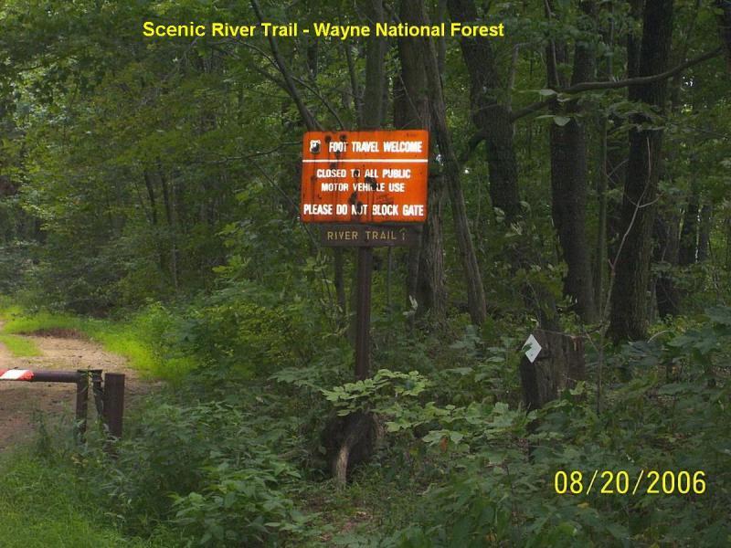 A wooden sign at the entrance of the Scenic River Trail in Wayne National Forest, indicating that foot travel is welcome. The sign states that the trail is closed to all public motor vehicle use and requests that visitors do not block the gate. The surrounding area features lush greenery and a dirt pathway leading into the forest. Scenic River Trail mountain bike trail.