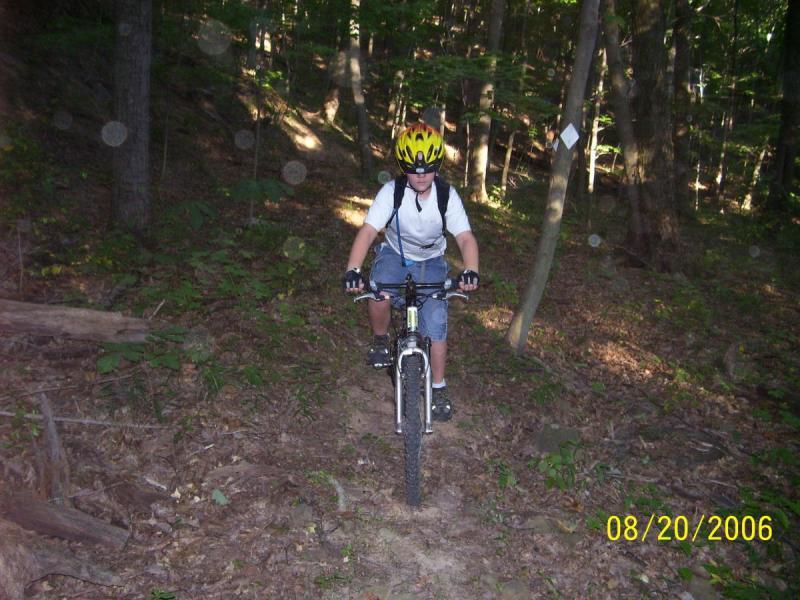 A child wearing a bright yellow helmet and sunglasses rides a mountain bike along a dirt trail in a forest. The trail is surrounded by trees and greenery, and the sun filters through the leaves, creating a dappled light effect. The child is dressed in a white t-shirt, denim shorts, and black gloves, focused on navigating the path ahead. The image is dated August 20, 2006. Scenic River Trail mountain bike trail.