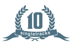 A circular logo featuring a laurel wreath, with the number "10" prominently displayed in the center. Below, the word "singletracks" is written in a bold, modern font. The color scheme is a muted blue, giving the design a clean and contemporary look.