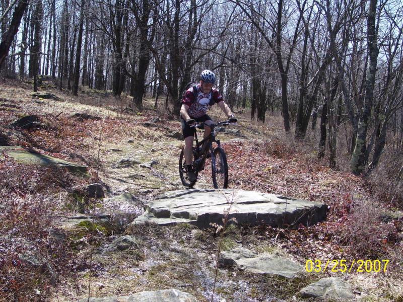 A cyclist navigating a rocky, outdoor mountain biking trail surrounded by bare trees and sparse vegetation. The image was taken on March 25, 2007. CVI Trails mountain bike trail.