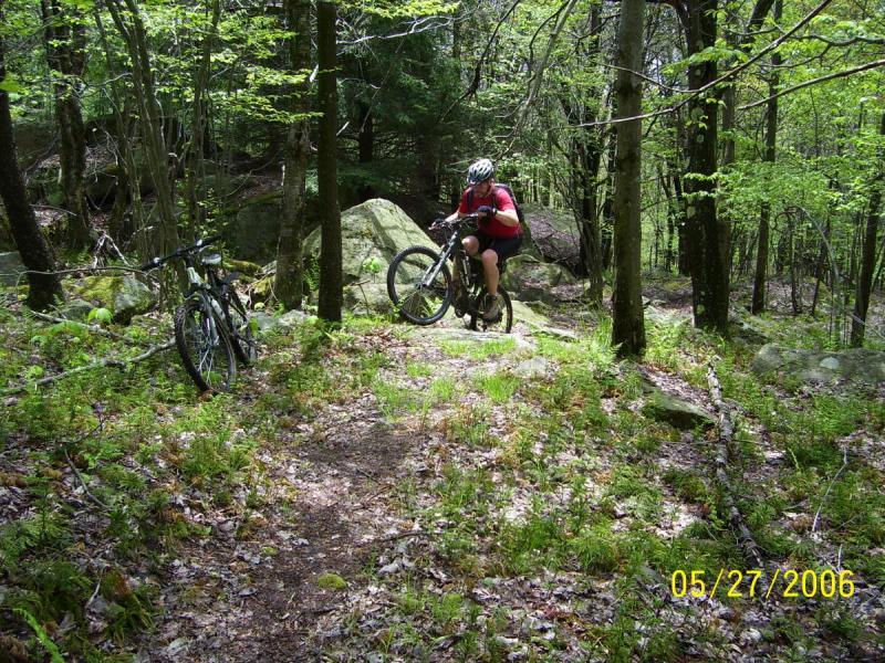 A mountain biker navigating a rocky path in a lush green forest, with two bikes parked nearby. The scene captures a bright, sunny day with trees and foliage providing a vibrant backdrop. CVI Trails mountain bike trail.