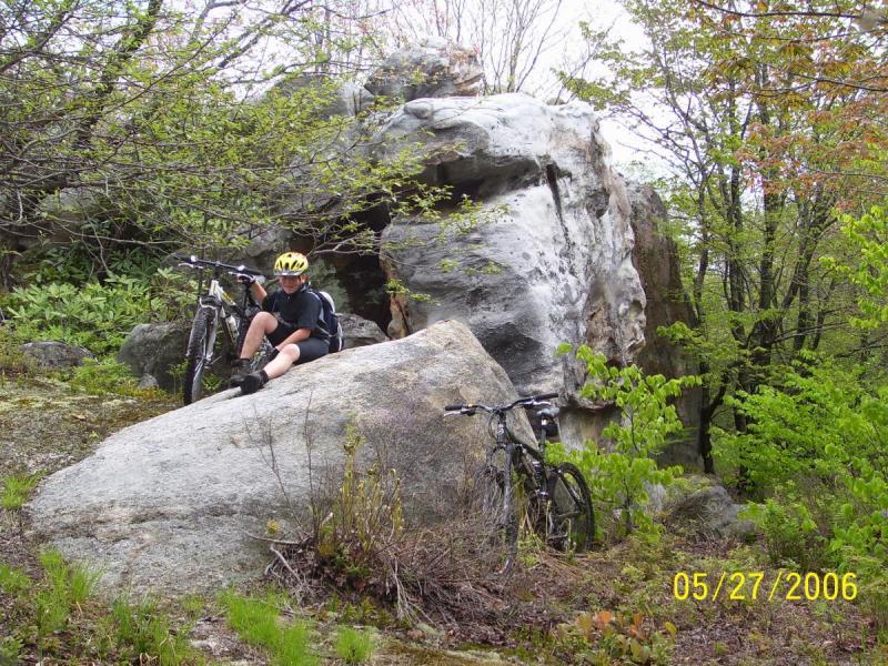 A young person wearing a yellow helmet sits on a large rock, taking a break from mountain biking in a lush, green forested area. Two bicycles are parked nearby, surrounded by greenery and natural rock formations. CVI Trails mountain bike trail.